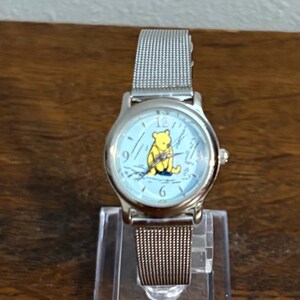 May include: Silver-toned wristwatch with a light blue face featuring a yellow Winnie the Pooh graphic. The watch has a mesh band and is displayed on a clear stand. A classic timepiece.