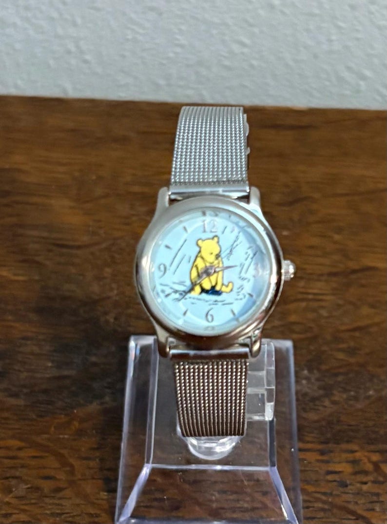 May include: Silver-toned wristwatch with a light blue face featuring a yellow Winnie the Pooh graphic. The watch has a mesh band and is displayed on a clear stand. A classic timepiece.
