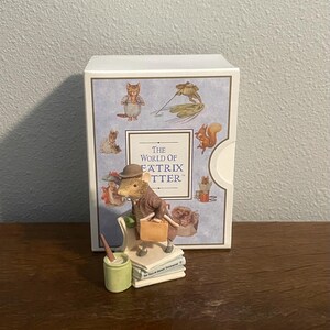 Peut inclure: Figurine d'une souris en costume et chapeau marron, tenant une mallette, assise sur des livres, et à côté d'un pot de peinture vert. La figurine est devant une boîte avec des illustrations de personnages animaux et le texte "The World of Beatrix Potter."