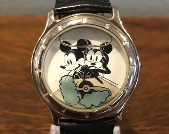 Könnte beinhalten: Eine silberfarbene Armbanduhr mit einem schwarzen Lederarmband. Das Zifferblatt der Uhr zeigt ein Cartoon-Bild von Mickey Mouse und Minnie Mouse, die ein Auto fahren.