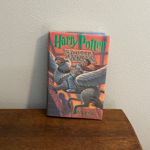 May include: A book cover for "Harry Potter and the Prisoner of Azkaban" by J.K. Rowling. The cover features a dark gray creature with wings, a young boy riding on its back, and the text "Harry Potter" in green letters.