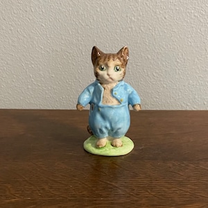May include: A small porcelain figurine of a brown and white cat wearing a blue jumpsuit. The cat is standing on a green base.