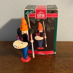 May include: A Hallmark Keepsake ornament of a nutcracker artist. The artist wears a purple jacket, blue pants, and a red-orange hat with red dots. He holds a paintbrush and a palette with colorful paint. The box reads "Franz The Artist."