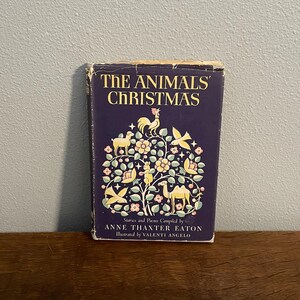 Op de afbeelding: Een vintage boek getiteld "The Animals' Christmas" met een donkerblauwe omslag. De omslag toont een kleurrijke illustratie van dieren, bloemen en vogels. Het boek is samengesteld door Anne Thaxter Eaton en geïllustreerd door Valenti Angelo.