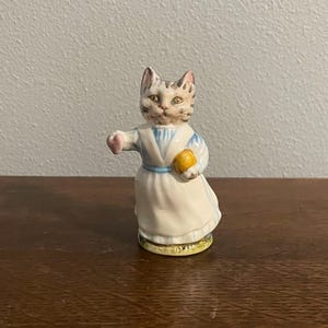 Puede incluir: Una figurita de cerámica de un gato con un vestido blanco con detalles azules y sosteniendo una pelota amarilla. El gato tiene pelaje marrón y blanco y ojos verdes.