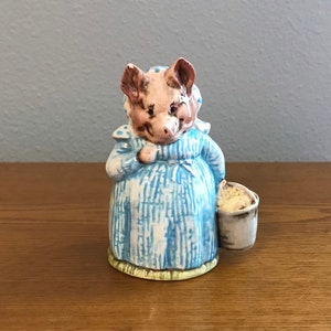 May include: A ceramic figurine of a pig wearing a blue and white dress and holding a bucket. The pig has a pink face and a white snout.