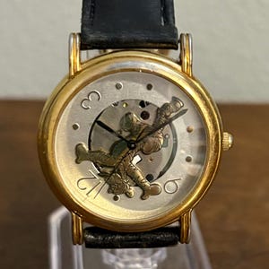 Könnte beinhalten: Goldfarbene Armbanduhr mit schwarzem Lederarmband. Das Zifferblatt hat einen silbernen Hintergrund mit goldenen römischen Ziffern und einem detaillierten Golfmotiv. Die Uhr hat einen klassischen, eleganten Stil.