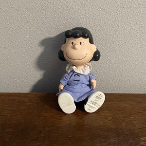 Könnte beinhalten: Eine Figur der Figur Lucy aus dem Peanuts-Comic. Lucy wird mit schwarzen Haaren, einem blauen Kleid und weißen Schuhen dargestellt. Die Figur sitzt mit einem lächelnden Gesichtsausdruck.