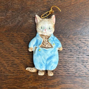 Può includere: Statuina di gatto in ceramica con un completo azzurro chiaro con bottoni dorati. Il gatto ha un muso marrone e bianco con occhi verdi e un filo d'oro per appenderlo. La statuina è su una superficie di legno.