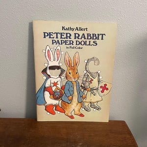 May include: A vintage book titled "Peter Rabbit Paper Dolls in Full Color" by Kathy Allert. The cover features illustrations of Peter Rabbit and two other rabbit paper dolls in costumes. The book is on a wooden surface.