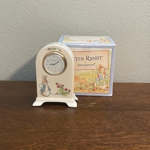 Puede incluir: Un reloj Peter Rabbit crema de Wedgwood. El reloj tiene una esfera redonda con números y manecillas negras. La base del reloj presenta una ilustración de Peter Rabbit. Una caja a juego está detrás del reloj.
