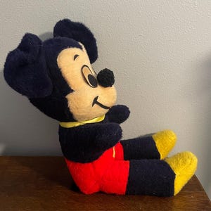 May include: A vintage Mickey Mouse plush toy, featuring a black body, ears, and arms, with a tan face and yellow feet. The plush is wearing red shorts and a yellow collar. The toy is sitting on a wooden surface.