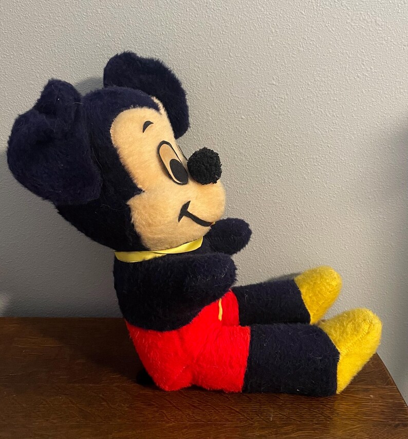 May include: A vintage Mickey Mouse plush toy, featuring a black body, ears, and arms, with a tan face and yellow feet. The plush is wearing red shorts and a yellow collar. The toy is sitting on a wooden surface.