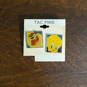 May include: A set of two enamel pins featuring cartoon characters, Tweety Bird and Taz. The pins are packaged on a white card with the text "TAC PINS" printed on it.