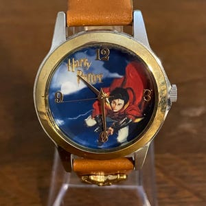May include: A wristwatch with a gold-colored bezel and a brown leather band. The watch face features the words "Harry Potter" and an image of a person flying on a broomstick against a blue sky. The watch has a silver-colored case.