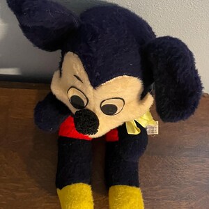 May include: Vintage Mickey Mouse plush toy. The plush features a cream-colored face, large black ears, and black body with red shorts and yellow feet. A yellow ribbon is tied around the neck. The toy is a classic Disney collectible.