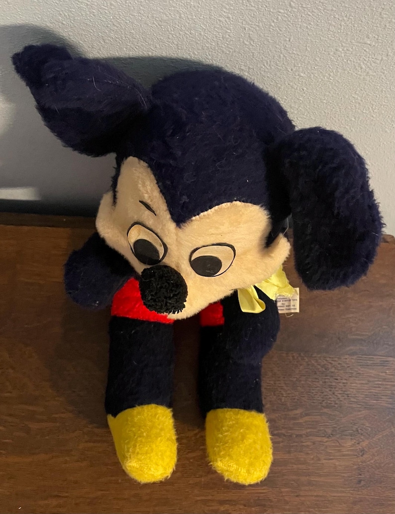 May include: Vintage Mickey Mouse plush toy. The plush features a cream-colored face, large black ears, and black body with red shorts and yellow feet. A yellow ribbon is tied around the neck. The toy is a classic Disney collectible.