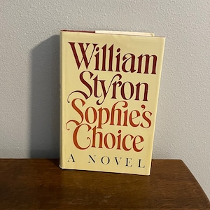 May include: A vintage paperback book titled "Sophie's Choice" by William Styron. The book cover is cream colored with red lettering. The text "A Novel" is printed in smaller red letters below the title.