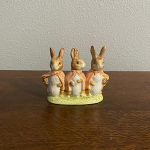 May include: A ceramic figurine featuring three rabbits wearing pink capes. The rabbits stand on a green base, with their fur in shades of brown and white. The figurine is likely a decorative item.