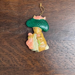 Può includere: Un ornamento in ceramica di Peter Rabbit con un cappello verde e un vestito rosa. L'ornamento è marrone e ha una corda dorata per appenderlo.