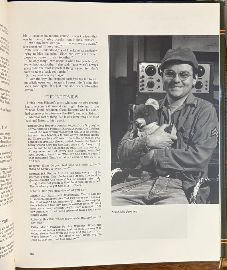 Op de afbeelding: Een zwart-witfoto van een man in militair uniform, met een bril en een pet, die een teddybeer vasthoudt. De man glimlacht. De afbeelding komt uit een boek, met tekst op de omliggende pagina's.