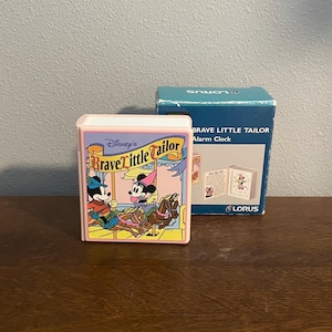 May include: A vintage Disney "Brave Little Tailor" alarm clock. The clock has a pink frame with a colorful illustration of Mickey and Minnie Mouse on a carousel. The box is blue and reads "Brave Little Tailor Alarm Clock".