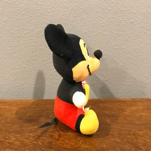 May include: A plush Mickey Mouse toy, with black ears, head, and a yellow face. The body is black, with red shorts and yellow feet. A small pink bow is around the neck. The toy is positioned on a wooden surface.