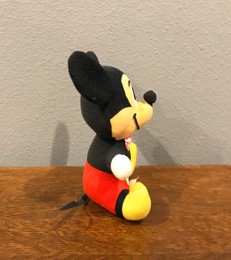 May include: A plush Mickey Mouse toy, with black ears, head, and a yellow face. The body is black, with red shorts and yellow feet. A small pink bow is around the neck. The toy is positioned on a wooden surface.