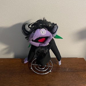 May include: A plush toy of a purple vampire-like character with black hair, a black suit, and a red mouth. The character has one visible eye and green accents. It is sitting on a metal stand.
