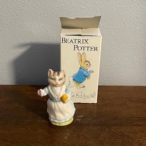 Puede incluir: Figura de cerámica de un gato con vestido, sosteniendo una bola amarilla, con una caja que presenta un conejo con chaqueta azul. La caja dice "Beatrix Potter" y "Beswick."