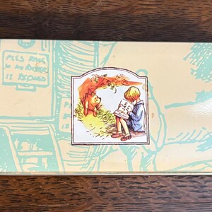 May include: Rectangular tin with a light yellow background and a Winnie the Pooh illustration. The illustration shows Pooh Bear and Christopher Robin reading. The tin has a light blue design of a door and stairs.