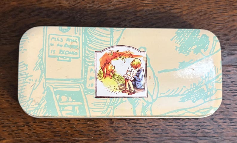 May include: Rectangular tin with a light yellow background and a Winnie the Pooh illustration. The illustration shows Pooh Bear and Christopher Robin reading. The tin has a light blue design of a door and stairs.