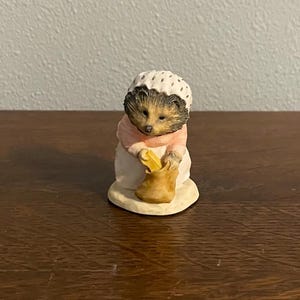 Puede incluir: Una pequeña figurita de cerámica de un erizo con un gorro blanco con lunares negros y un vestido rosa. El erizo sostiene una bolsa marrón y una hogaza de pan amarilla.