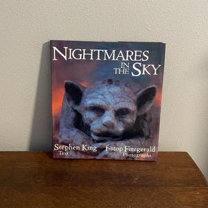 Könnte beinhalten: Ein Buch mit dem Titel "Nightmares in the Sky" von Stephen King mit Fotografien von f-stop Fitzgerald. Das Cover zeigt eine Nahaufnahme einer Gargoyle-Skulptur vor einem dunklen, stürmischen Himmel.