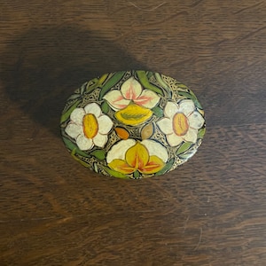 May include: A round, wooden trinket box with a floral design. The box is painted with white, yellow, and orange flowers on a dark green background.