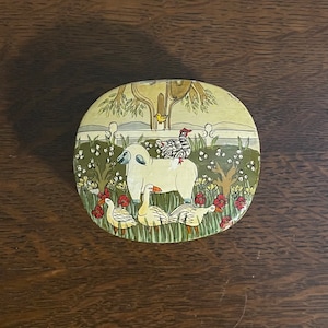 May include: A round, painted tin box with a white sheep, a black and white hen, and three yellow ducks in a green field with red flowers. The box has a white fence and a yellow bird in a tree.