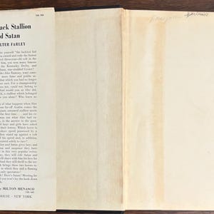 May include: Open book with the title "The Black Stallion and Satan" by Walter Farley. The book's pages are off-white with black text. The cover is black. The book is illustrated by Milton Menasco and published by Random House, New York.