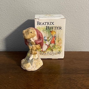 Könnte beinhalten: Eine Keramikfigur von Mr. Toad aus The Wind in the Willows von Beatrix Potter. Mr. Toad trägt eine rote Jacke und hält eine Schaufel. Die Figur steht auf einem Sockel mit braunem und grünem Design. Die Figur ist in einer Schachtel mit dem Beatrix Potter Logo und John Beswick verpackt.