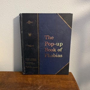 May include: A hardcover book titled "The Pop-up Book of Phobias" with a navy blue cover and gold lettering. The spine is black with gold accents and illustrations. The book is on a wooden surface.