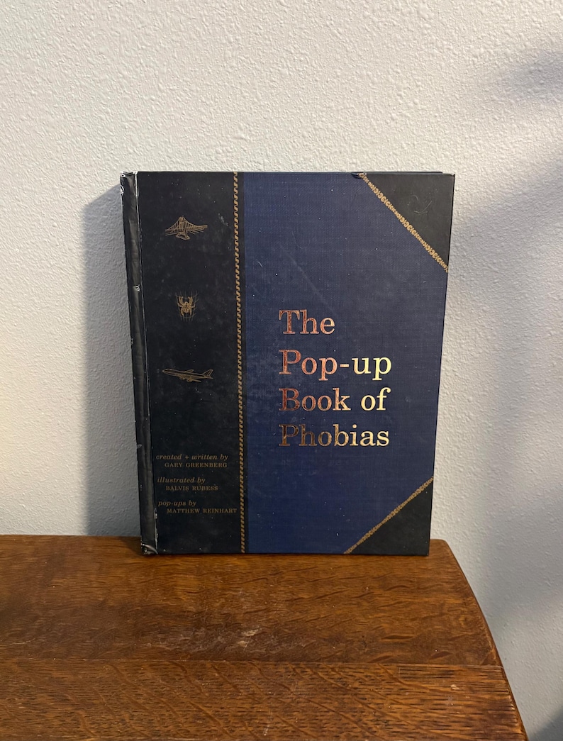 May include: A hardcover book titled "The Pop-up Book of Phobias" with a navy blue cover and gold lettering. The spine is black with gold accents and illustrations. The book is on a wooden surface.