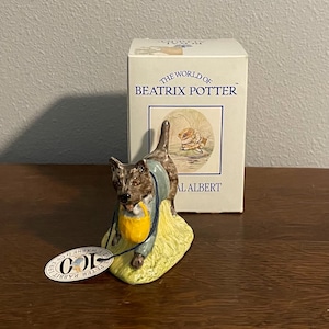 Puede incluir: Figura de cerámica de un perro con chaqueta azul y delantal amarillo, de pie sobre un trozo de hierba. La figura es parte de la colección "The World of Beatrix Potter". Una etiqueta con el texto "100" está adjunta.