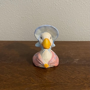 Pode incluir: Uma estatueta de pato de cerâmica usando um chapéu azul e um vestido rosa e branco. O pato tem um bico amarelo e uma pequena flor amarela no vestido. A estatueta está em uma superfície de madeira.