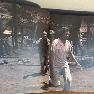 Op de afbeelding: Een zwart-witfoto toont drie mensen in een oorlogssituatie. E&eacute;n persoon staat op de voorgrond, gekleed in lichte kleding en rent. Twee anderen volgen, &eacute;&eacute;n met een helm en de ander met een hoed.