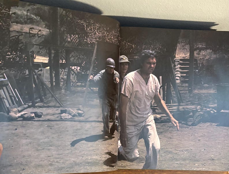 Op de afbeelding: Een zwart-witfoto toont drie mensen in een oorlogssituatie. E&eacute;n persoon staat op de voorgrond, gekleed in lichte kleding en rent. Twee anderen volgen, &eacute;&eacute;n met een helm en de ander met een hoed.