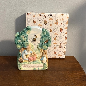 May include: A ceramic clock featuring a scene of four rabbits in a forest with green trees. The clock face has black numbers and gold hands. Text at the bottom reads: Once upon a time there were four little Rabbits, and their names were- Flopsy, Mopsy, Cotton-tail and Peter.