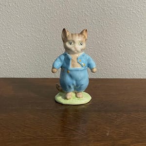 May include: A ceramic figurine of a cat wearing a light blue outfit. The cat has tan and brown fur and is standing on a green base. The figurine has a vintage aesthetic.