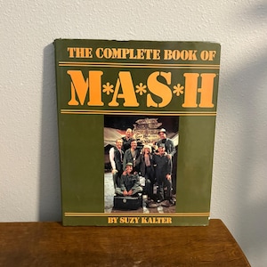Op de afbeelding: Een hardcover boek met de titel "THE COMPLETE BOOK OF M*A*S*H" met oranje letters op een groene achtergrond. De omslag toont een foto van een groep mensen. De naam van de auteur, SUZY KALTER, staat onderaan.