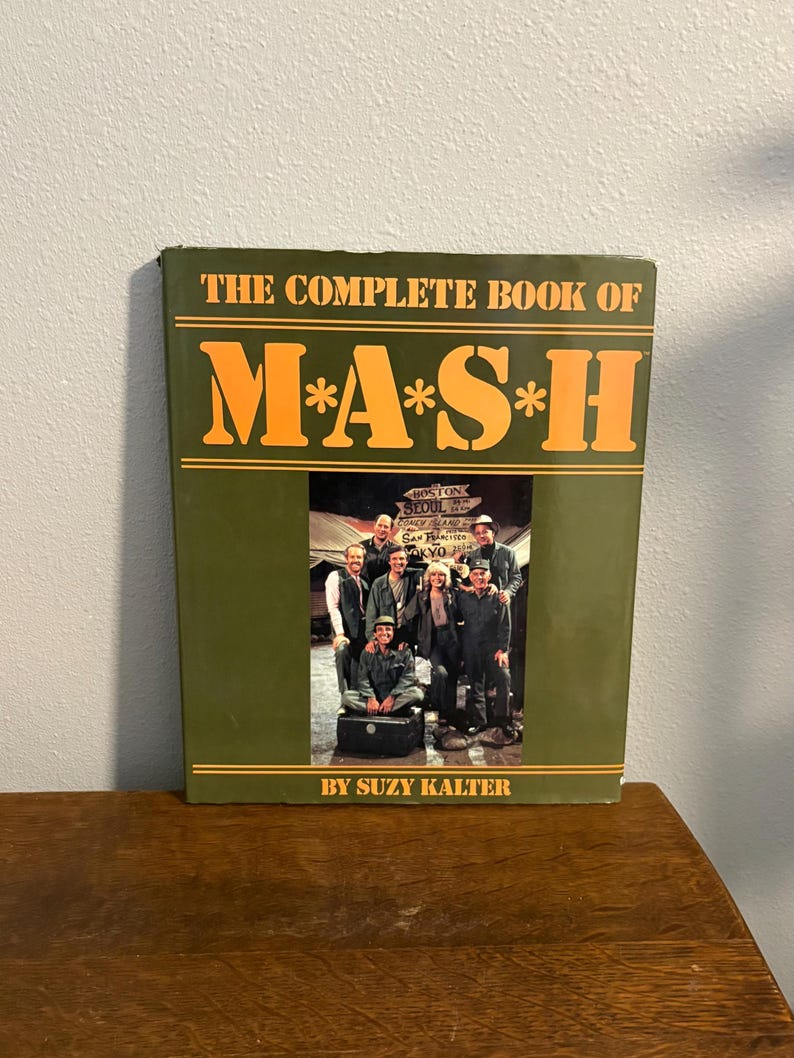 Op de afbeelding: Een hardcover boek met de titel "THE COMPLETE BOOK OF M*A*S*H" met oranje letters op een groene achtergrond. De omslag toont een foto van een groep mensen. De naam van de auteur, SUZY KALTER, staat onderaan.