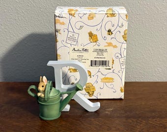 May include: A decorative Peter Rabbit figurine in a green watering can, with a white letter "R" and a patterned box in the background. The figurine features a small rabbit with a beige and white color scheme. The box has illustrations of Beatrix Potter characters.