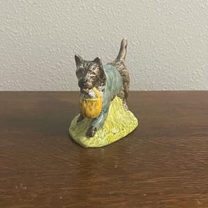 Puede incluir: Figura de cerámica de un perro que lleva una cesta amarilla. El perro es marrón y negro con una chaqueta azul claro. Se encuentra sobre un trozo de hierba verde. La figura está sobre una superficie marrón oscuro.